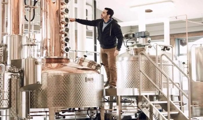 A person in a dark hoodie and brown pants adjusts settings on large copper and stainless steel distillation equipment inside an industrial facility.