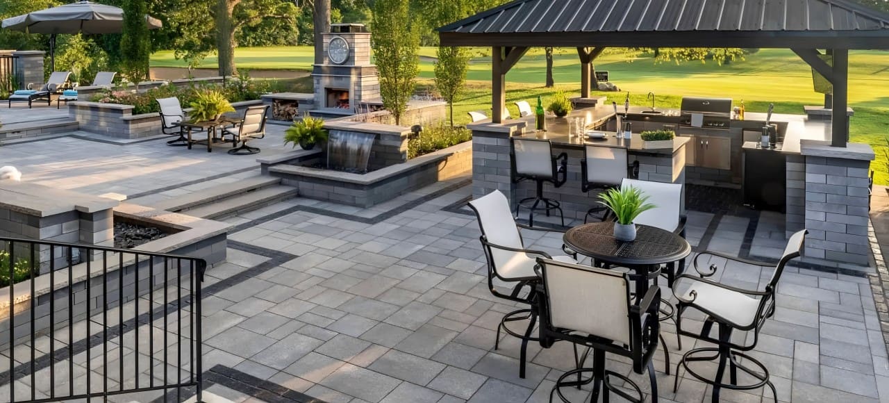 Outdoor patio with modern furniture, including a table with chairs, an outdoor kitchen under a gazebo, and a separate seating area with armchairs and a large clock on the wall, surrounded by greenery.