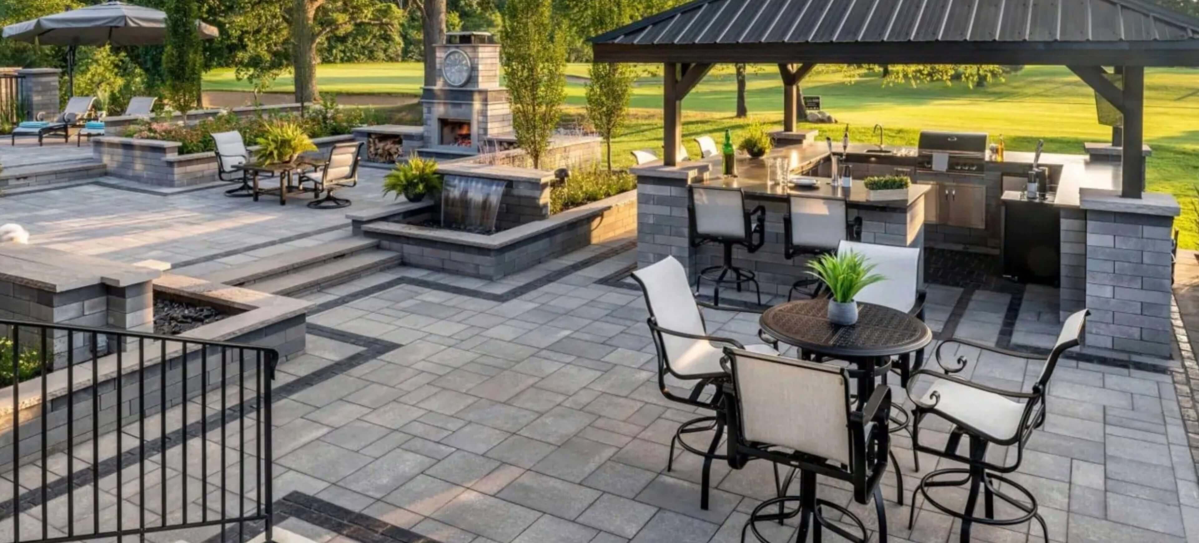A spacious outdoor patio with multiple seating areas, a wooden pergola, a fireplace, and a bar counter surrounded by greenery and a view of a park or garden.