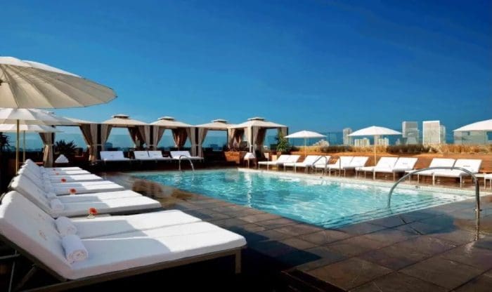 A rooftop pool with white lounge chairs and umbrellas arranged around it, cabanas in the background, and a cityscape visible in the distance under a clear blue sky.